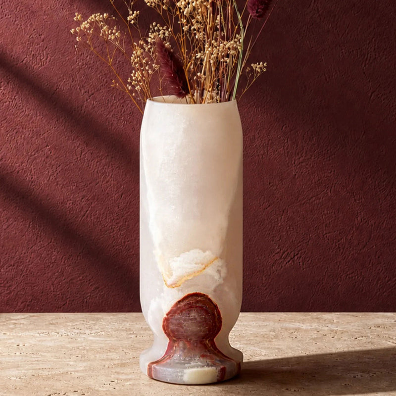 A tall cylindrical vase made of frosted off-white stone with a textured reddish-brown base, holding dried flowers, set on a light stone surface against a dark reddish-brown wall.