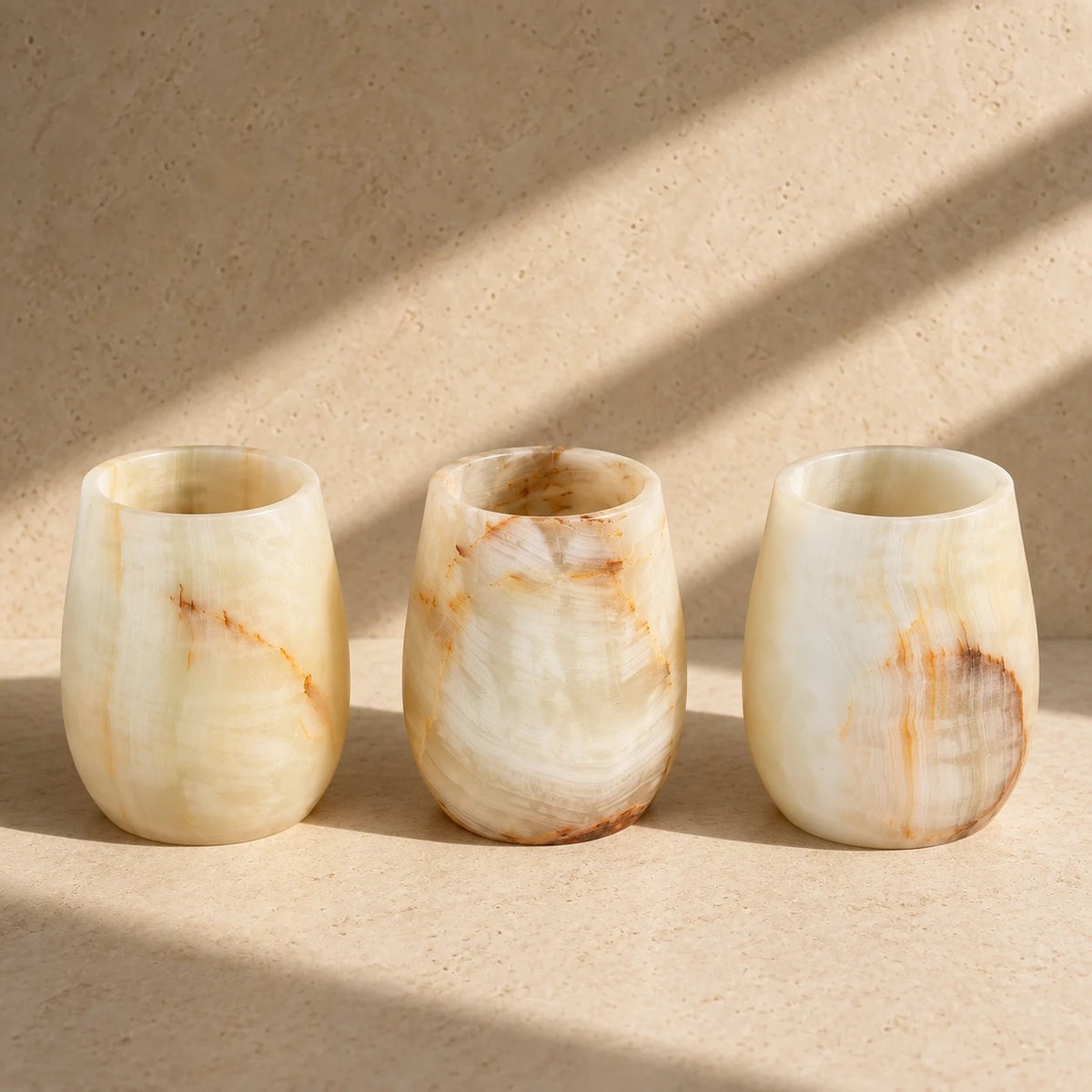 Three hand-carved ivory onyx curved vessels with honey and rust veining, side by side on a sandstone surface in slanted sunlight