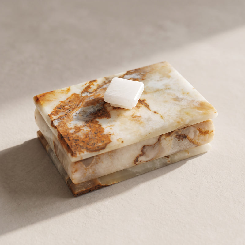 Ivory and rust onyx ring and trinket box with lid, seen from above at an angle: a creamy translucent lid with a rust and umber patch, a white square stone knob and a stepped base, on pale plaster in a slant of sunlight