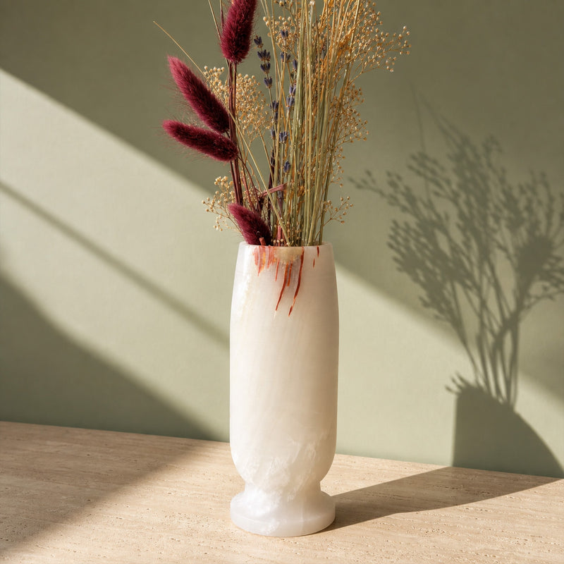 Ivory onyx vase with thin rust streaks running down from the rim and a honey patch at the lip, holding dried burgundy bunny tails, lavender and gypsophila on a travertine table against a sage-green wall