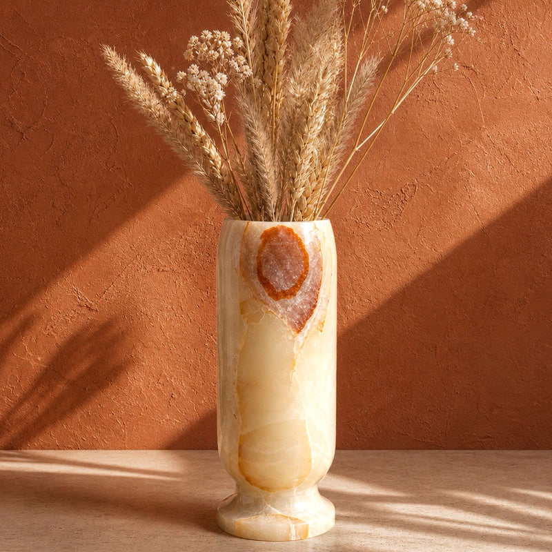 A cylindrical onyx stone vase with dried wheat and small white flowers, set on a light stone surface against a textured terracotta wall with warm sunlight and shadows.