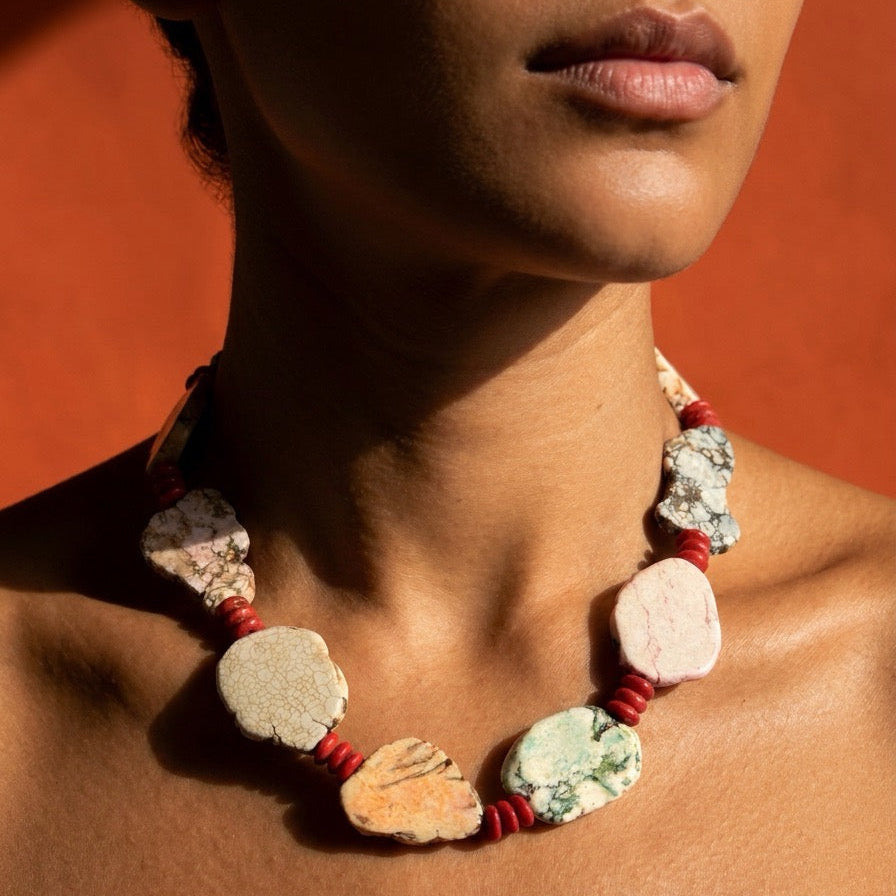 A close-up of a model wearing a chunky statement necklace made of irregular natural stone slices separated by red cord, set against a warm orange background.
