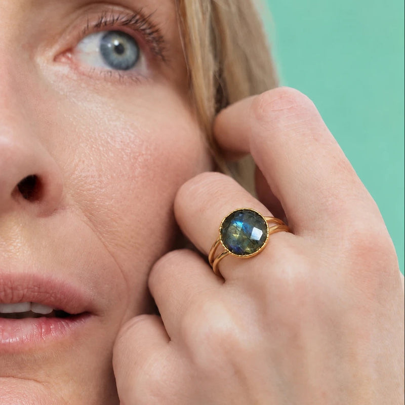 Faceted round labradorite ring worn on the hand, gray stone with a blue flash in a fine-edged 22k gold-filled bezel, fingers raised to the cheek against a mint green background
