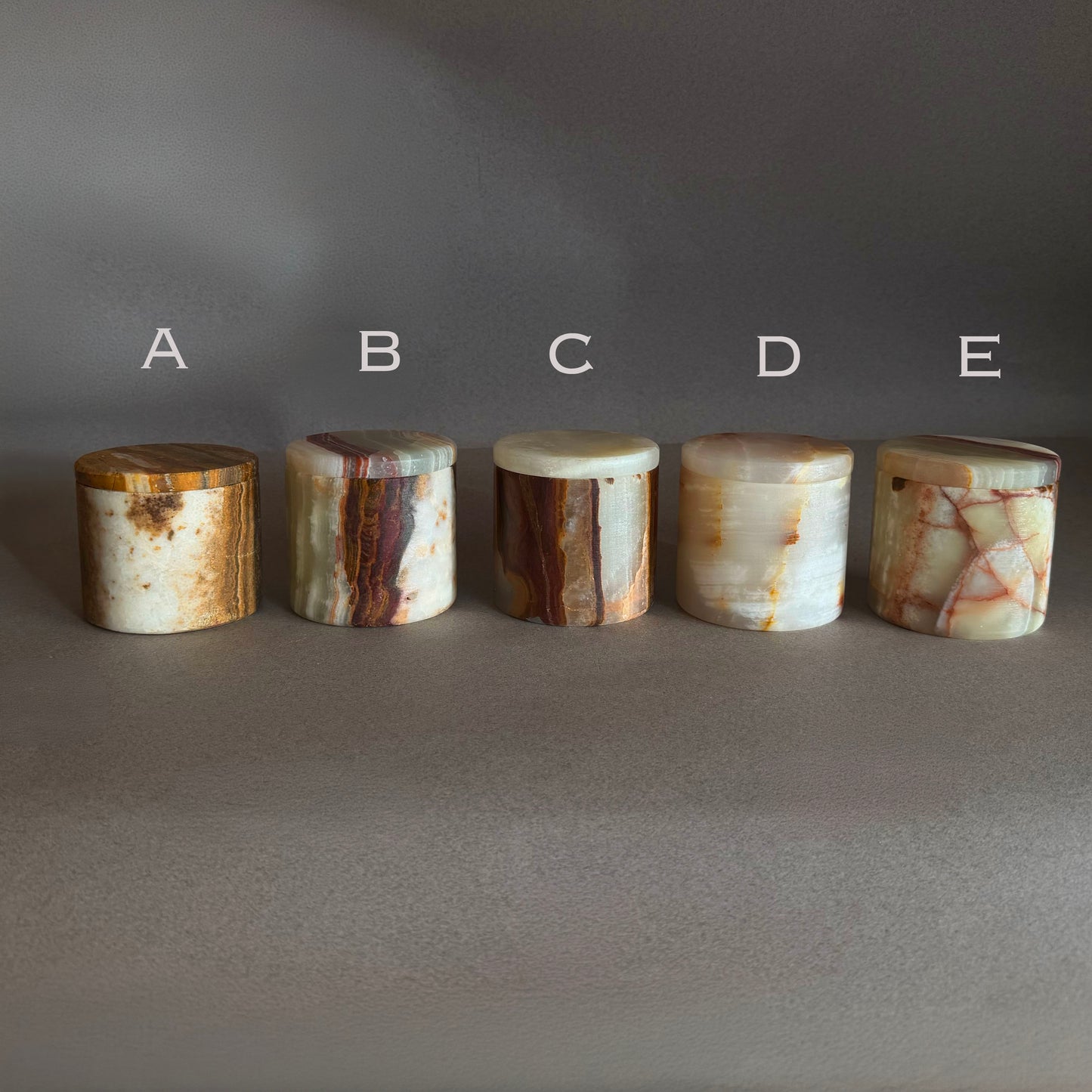 Five cylindrical onyx stone jars with lids salt jars labeled A to E on a gray background