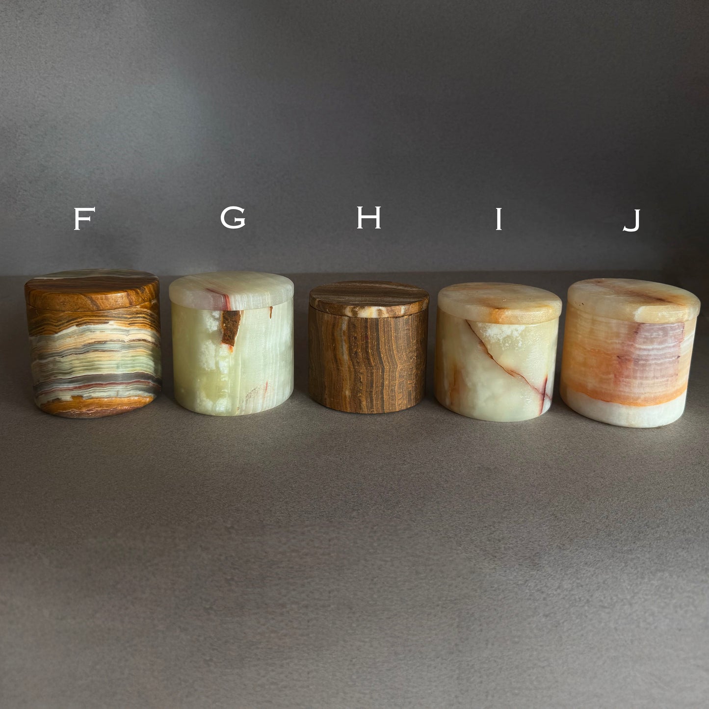 Five cylindrical onyx stone small jars with lids with different patterns arranged in a row on a gray surface.