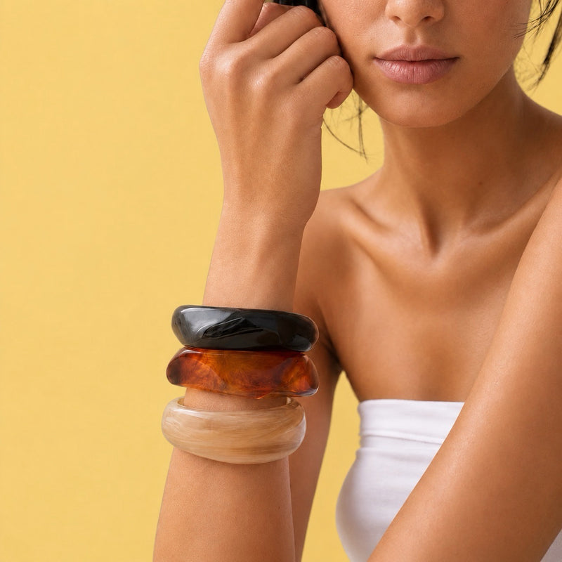 A close-up of a model's wrist wearing a stack of four smooth, polished stone-like bangles in black, amber-brown, translucent orange-brown, and light beige, set against a solid yellow background.
