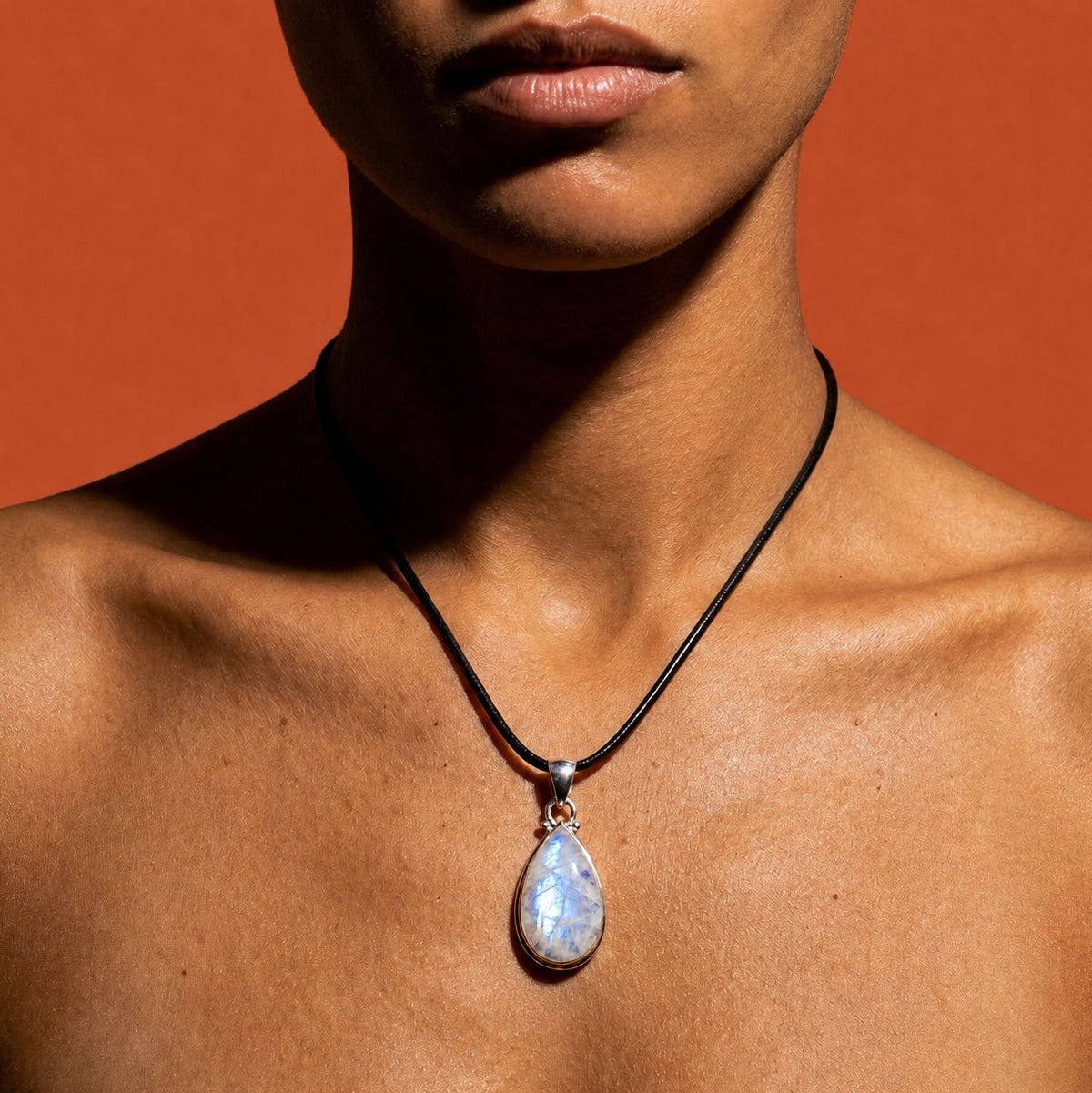 A close-up of a model wearing a black cord necklace with a teardrop-shaped iridescent moonstone pendant, set against a solid orange background.