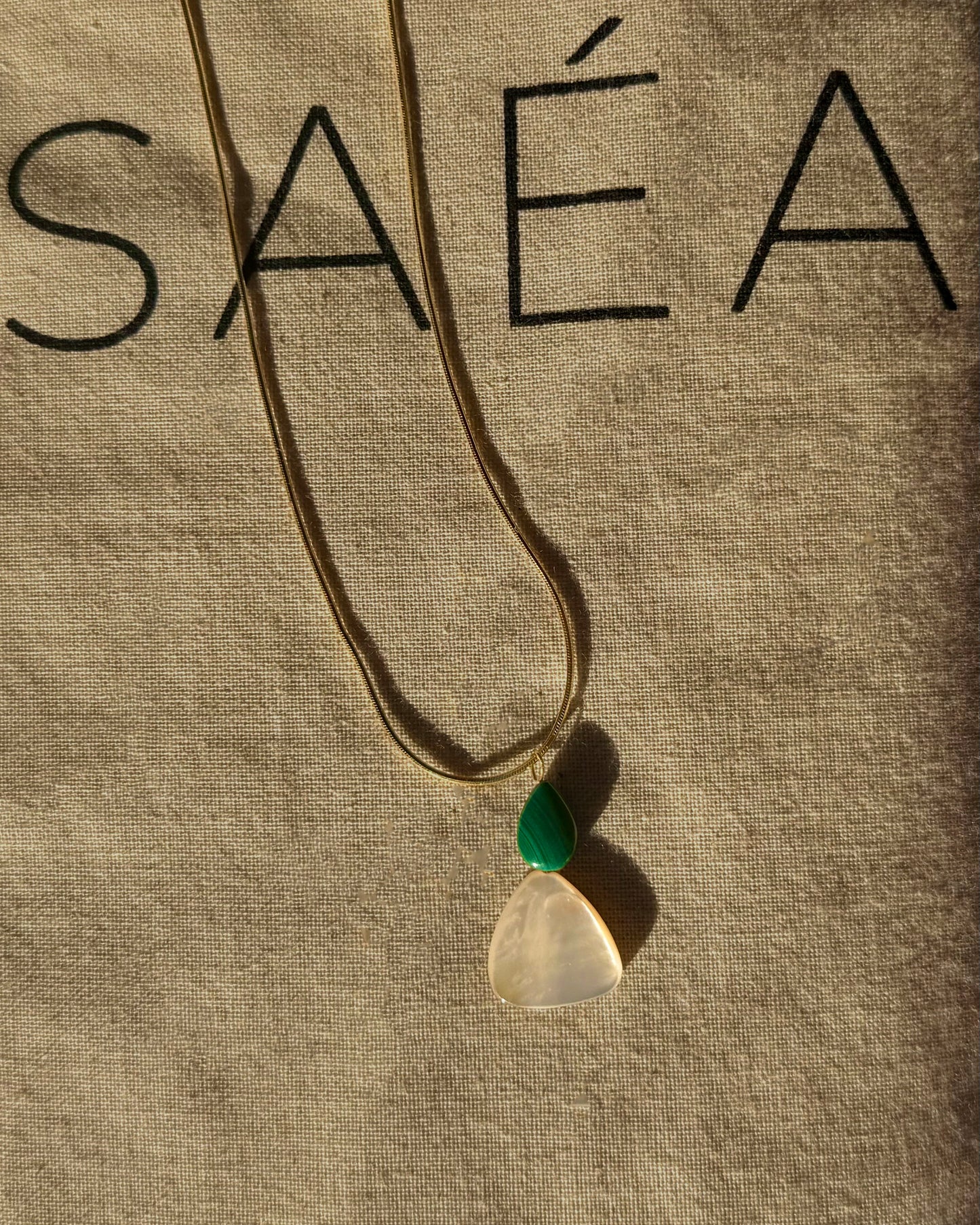 Gold Snake Chain Necklace with a pendant on a beige fabric background with SAÉA branding.