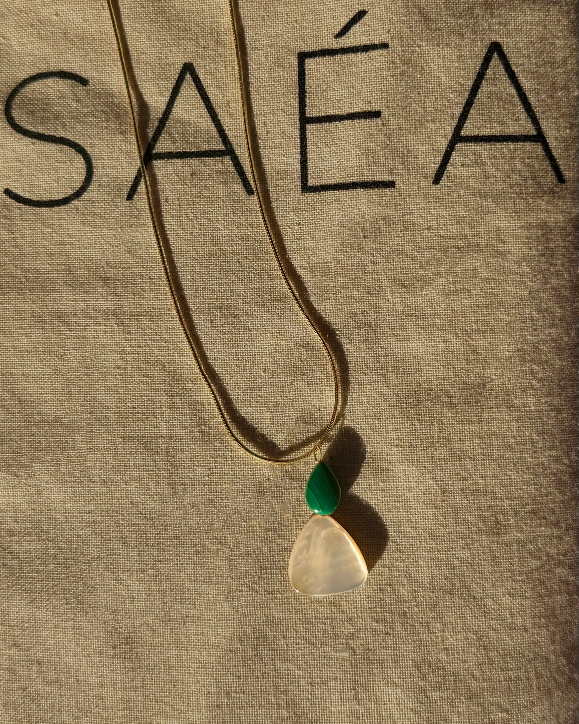 Gold Snake Chain Necklace with a pendant on a beige fabric background with SAÉA branding.