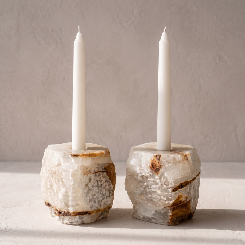 Two raw-edged natural onyx candle holders, one light and one darker, holding white taper candles on a pale plaster ledge.