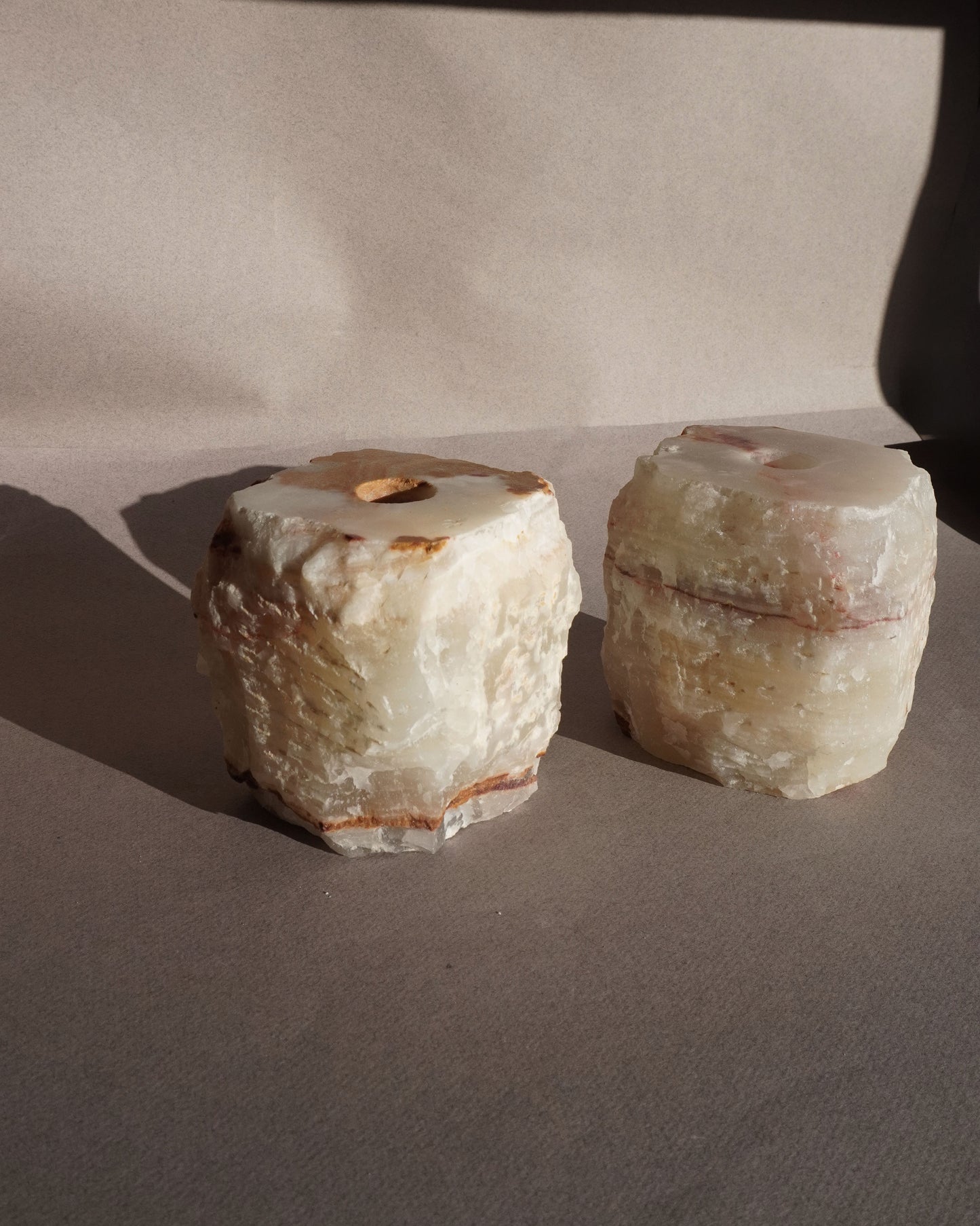 Two stone objects on a plain background Handmade onyx taper holders with cream and amber veining styled with white candles casting soft shadows
