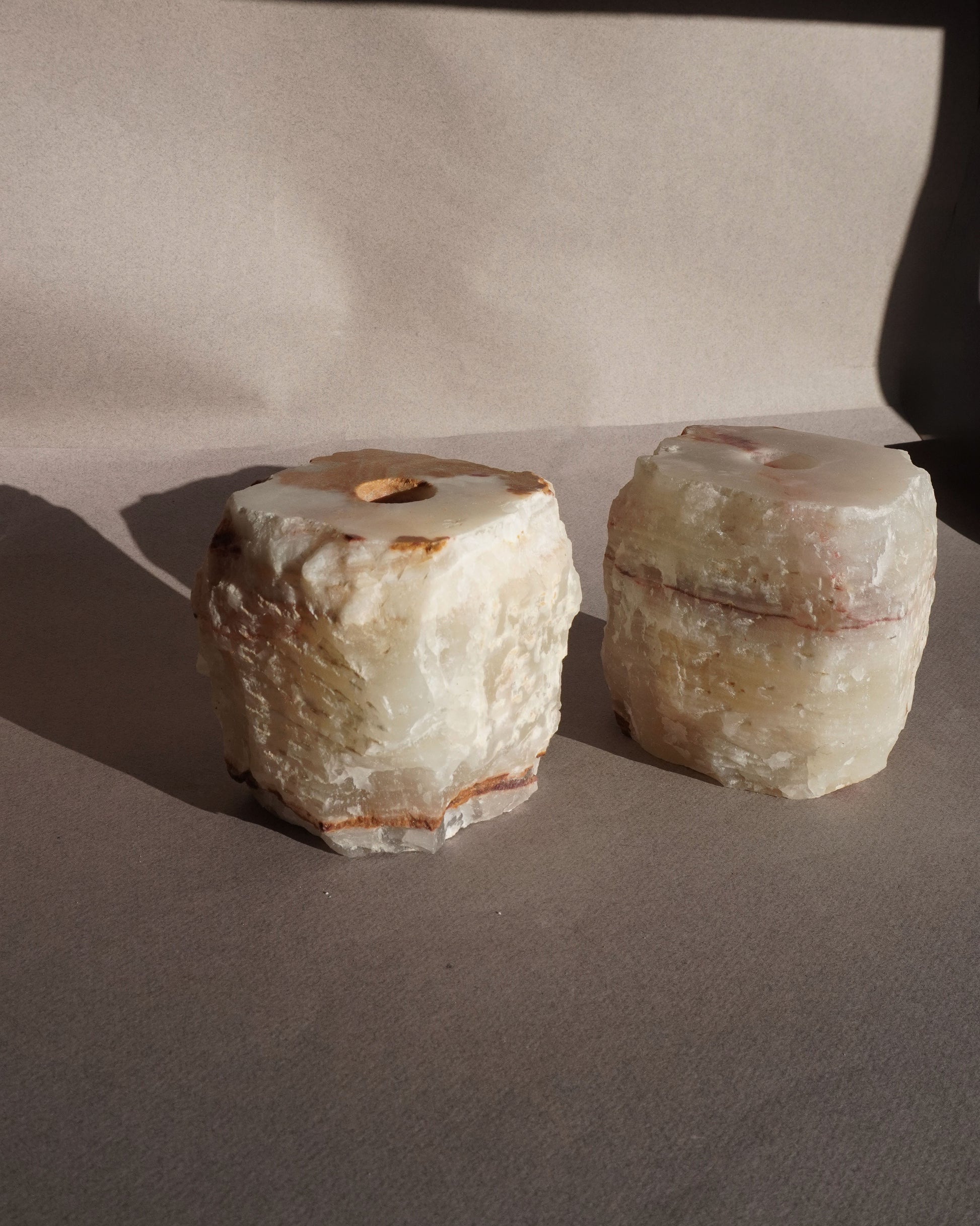Two stone objects on a plain background Handmade onyx taper holders with cream and amber veining styled with white candles casting soft shadows
