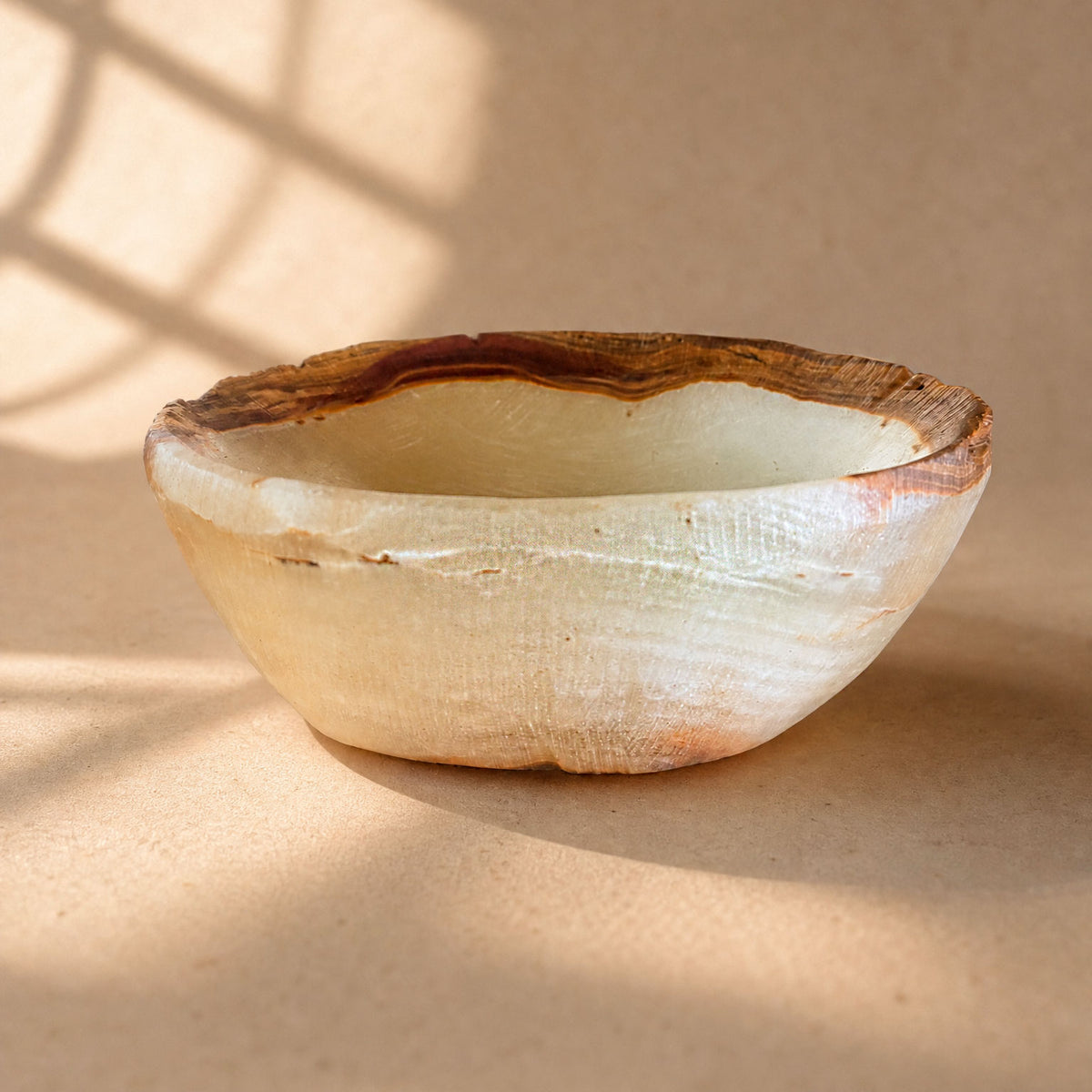Bone and burgundy raw edge onyx bowl, honey-brown striated rim with burgundy vein, on sunlit surface