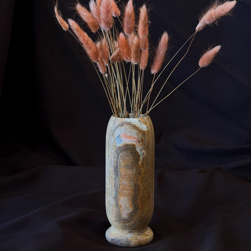 Onyx origin vase in ochre, smoke and blush with an amber banded core and a blush patch near the rim, holding pink bunny tail grass on dark fabric