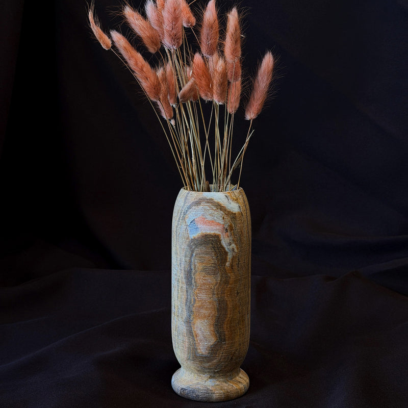 Onyx origin vase in ochre, smoke and blush with an amber banded core and a blush patch near the rim, holding pink bunny tail grass on dark fabric