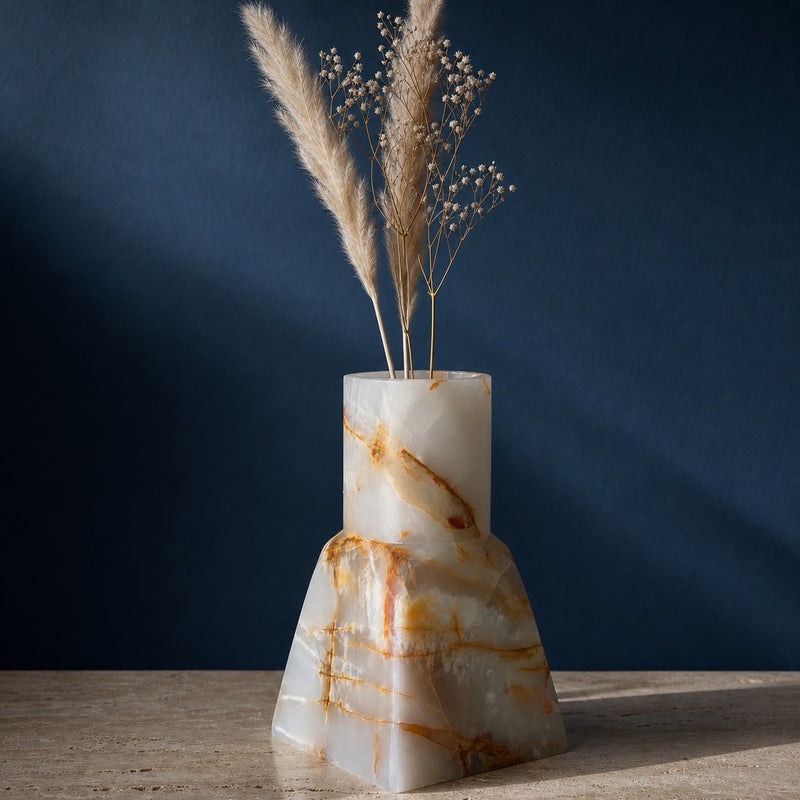 Hand-carved milk-white onyx bud vase with a straight neck on a tapered base, branching amber and gold veins, holding pampas and baby's breath against a deep navy wall