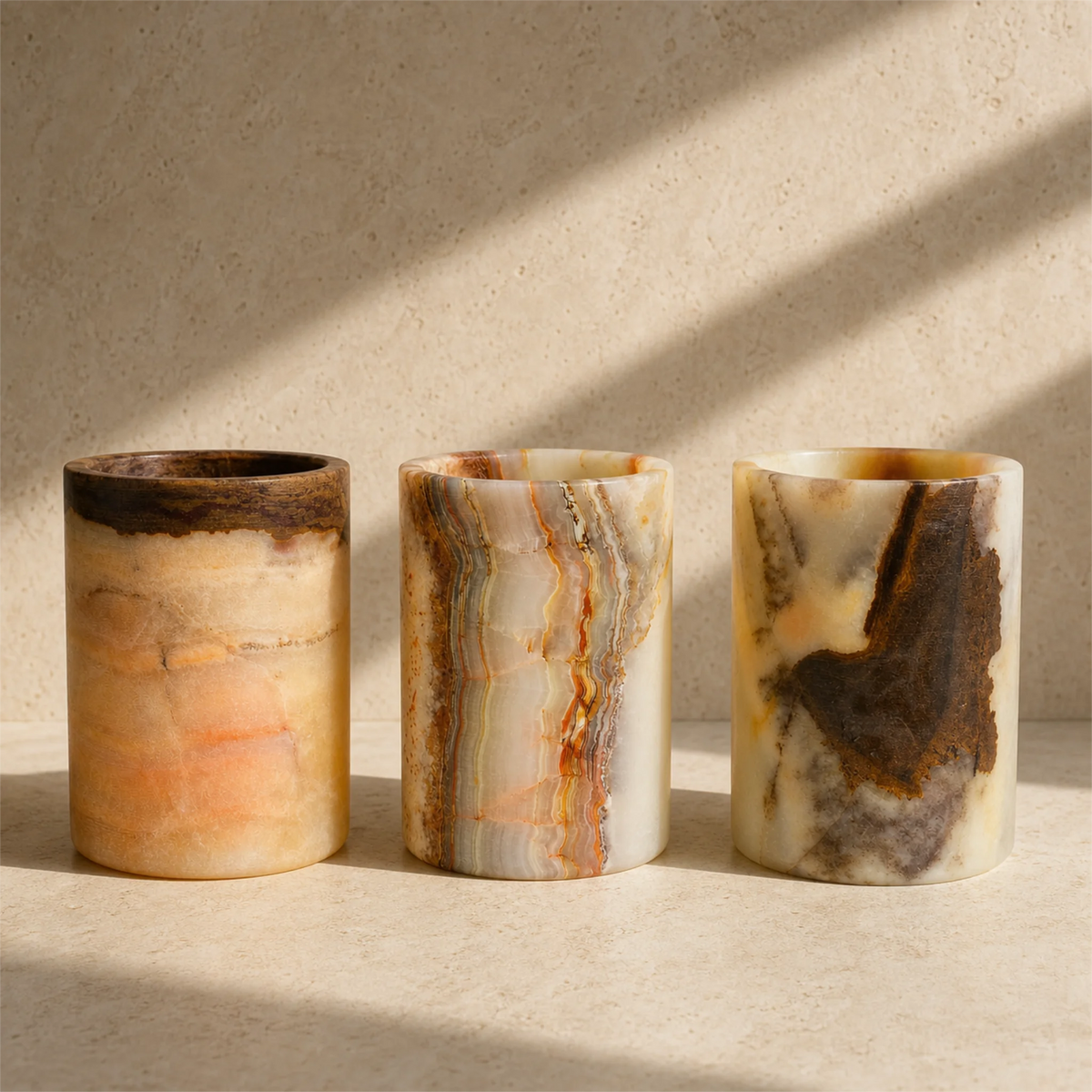 Three cylindrical stone toothbrush holders with natural marbled patterns in beige, cream, orange, and dark brown, arranged on a light stone surface with soft window shadows.