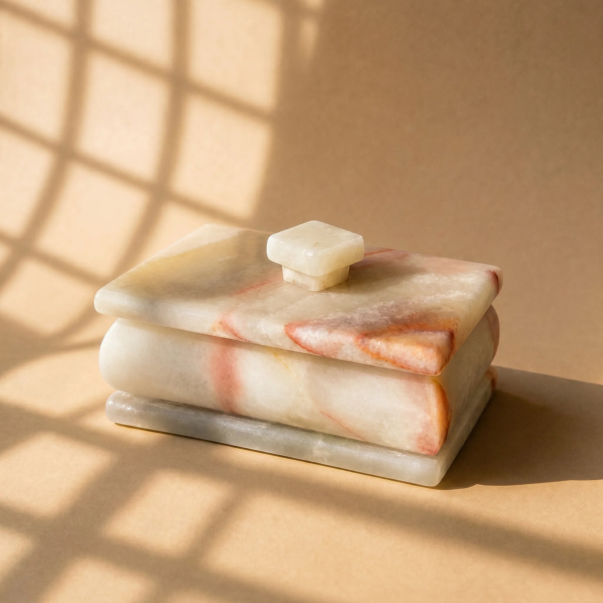 Angled view of ivory onyx lidded box with blush and coral veining and pale square knob in window light