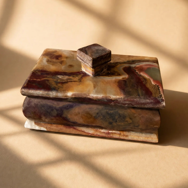 A small rectangular lidded box made of marbled stone in earthy brown, beige, and purple tones, photographed on a warm beige surface with soft window-shadow patterns cast across the scene.