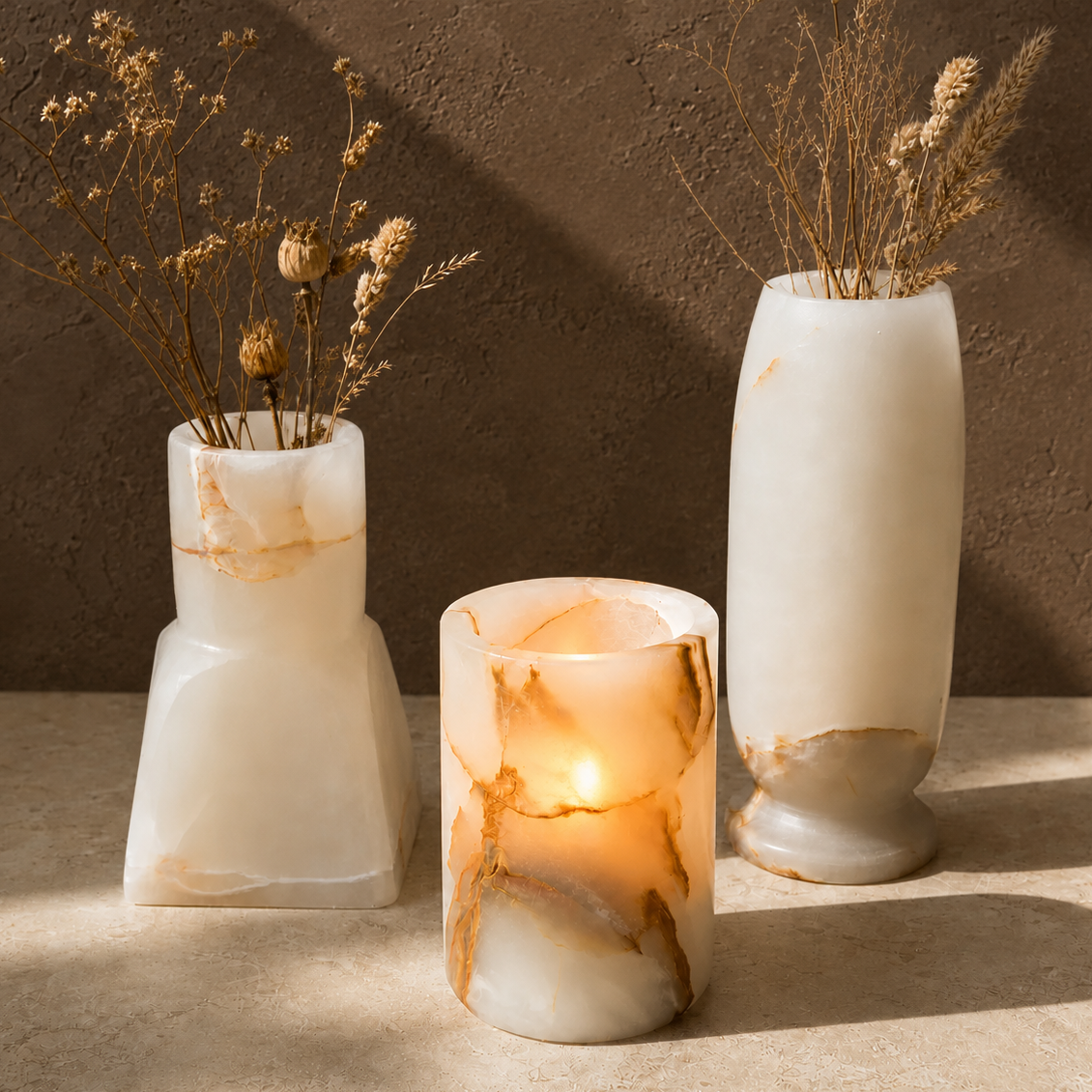 Three white onyx stone vases with dried floral arrangements, arranged on a beige surface against a textured brown wall. The center vase is lit from within, casting a warm glow.