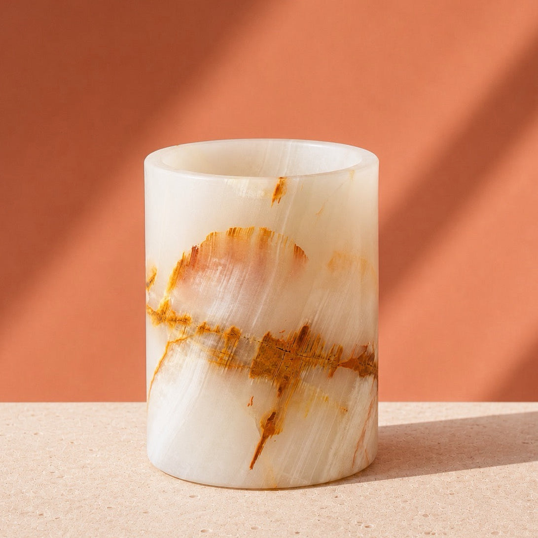 A cylindrical white onyx or marble candle holder with natural brown and orange veining, set on a textured beige surface against a warm terracotta background with diagonal shadows.