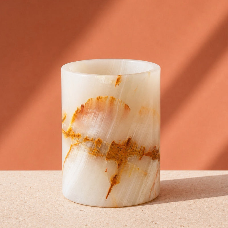 A cylindrical white onyx or marble candle holder with natural brown and orange veining, set on a textured beige surface against a warm terracotta background with diagonal shadows.