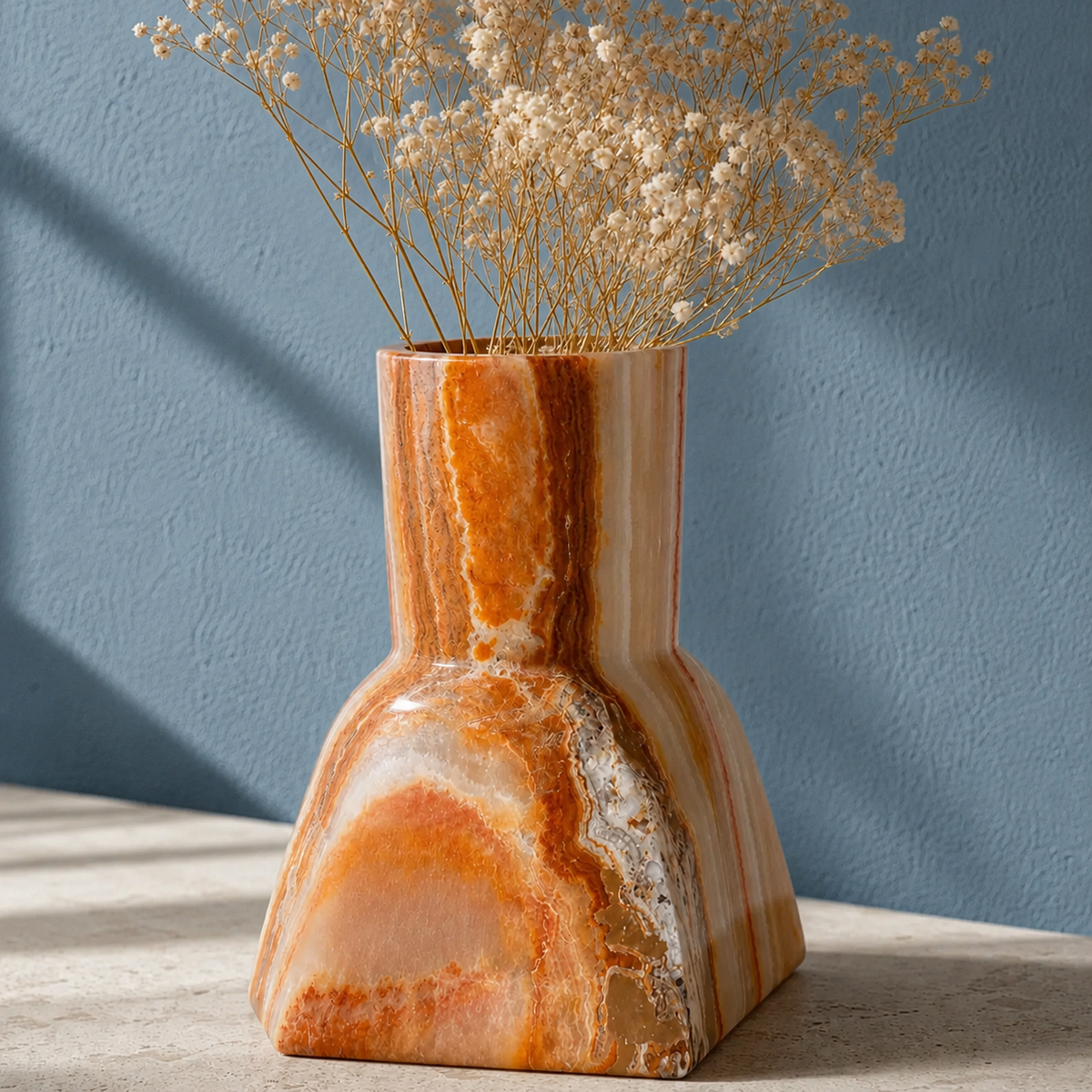A modern onyx rust vase made from polished orange and cream agate or onyx stone, holding dried baby's breath stems, set on a light stone surface against a textured blue wall.