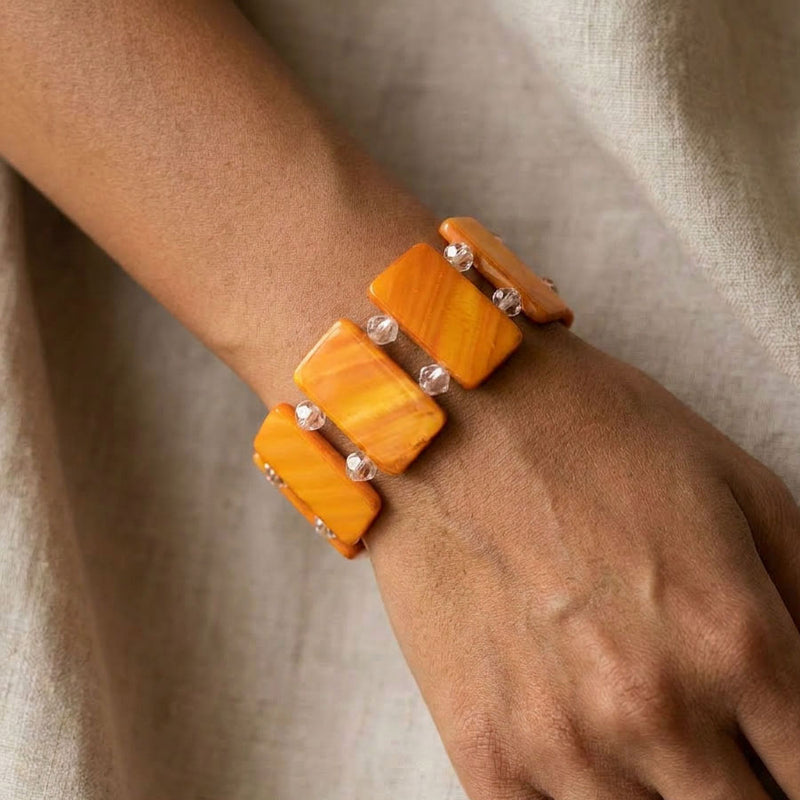 A close-up of a person's wrist and hand wearing a multi-strand bracelet made of warm orange-yellow rectangular stone-like pieces separated by small silver beads, set against a soft beige fabric background.

Orange dyed shell pearl stretch bracelet with faceted clear crystal spacers, worn on the wrist against neutral linen