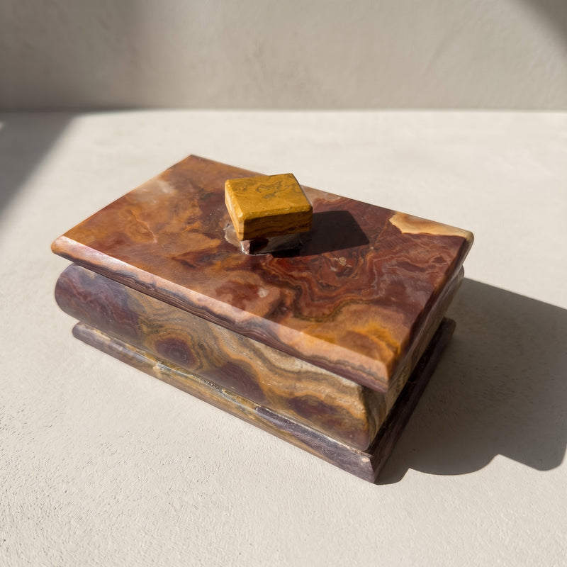 Rectangular onyx keepsake box with lid, three-quarter front view in raking sun on pale plaster, oxblood and caramel swirled lid over a plum body banded in gold, with a mustard stone knob