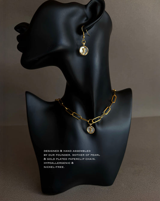 Gold chain paperclip necklace and mother of pearl earrings on a black mannequin bust with text on a gray background