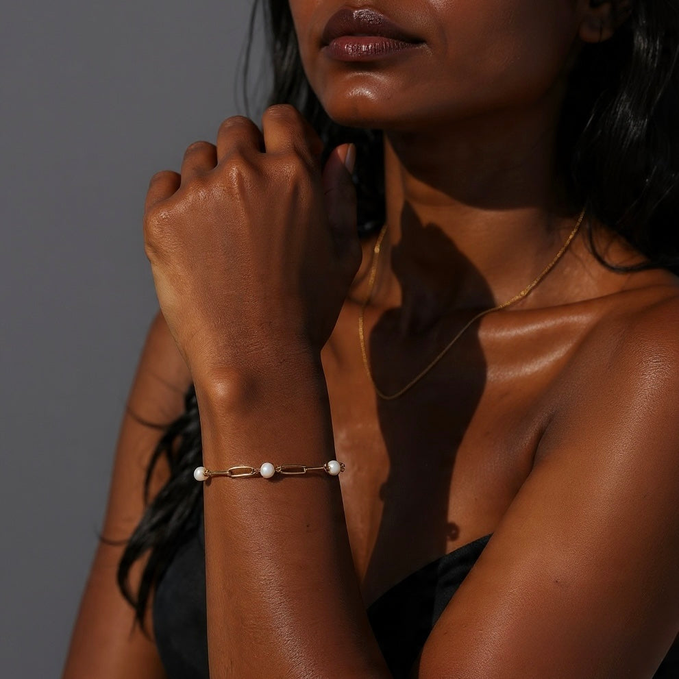 Gold paperclip chain bracelet with pearls shown on a woman’s wrist in natural sunlight.
 Pearl and gold paperclip bracelet styled on a woman with deep skin tone, photographed in warm sunlight.
