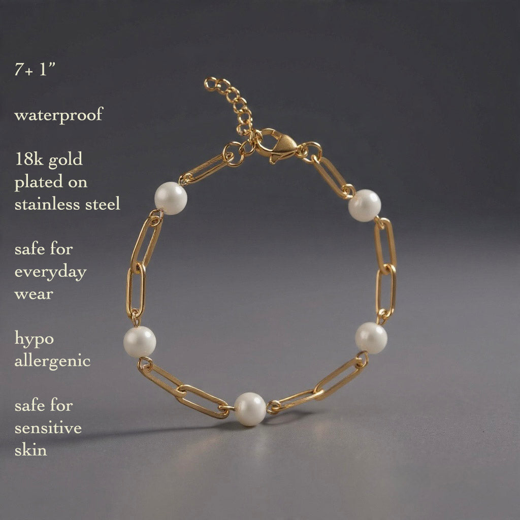 Close-up of gold paperclip chain bracelet with five round pearls on a gray background.
