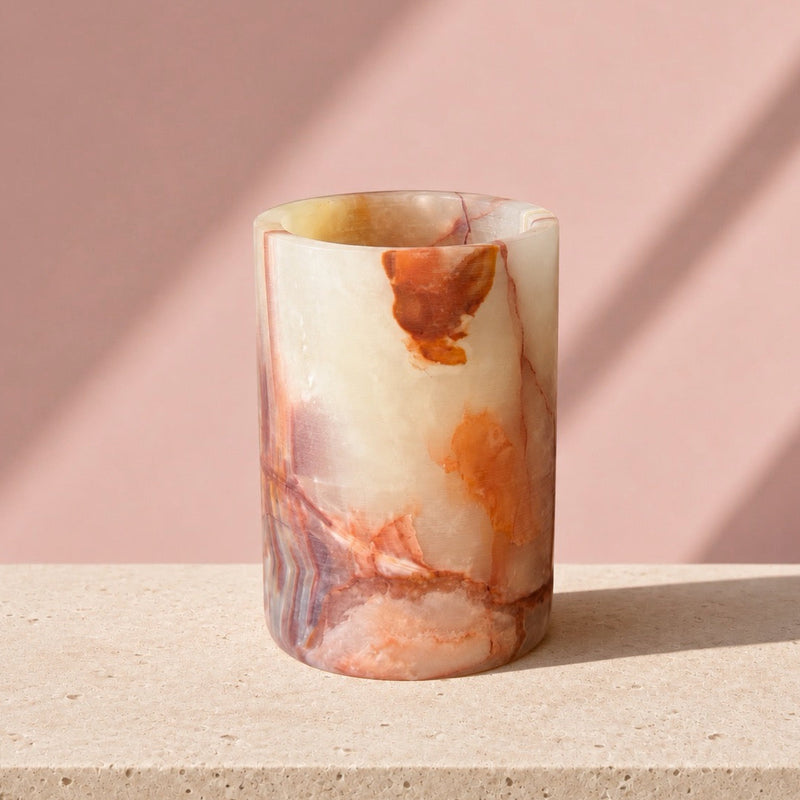 A cylindrical stone cup or vase made from polished onyx with swirling cream, orange, and reddish-brown marbling, standing on a light stone surface against a dusty pink background with soft diagonal shadows.