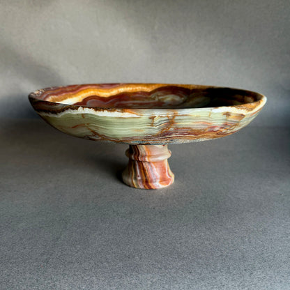 Translucent onyx pedestal bowl held against blue sky and palm trees, glowing olive green, amber, plum, and cream stone layers
