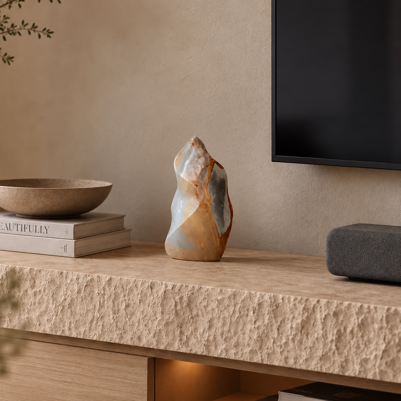 Hand-carved polychrome jasper flame sculpture in apricot, cream and pale gray standing on a stone media console beside books and a ceramic bowl