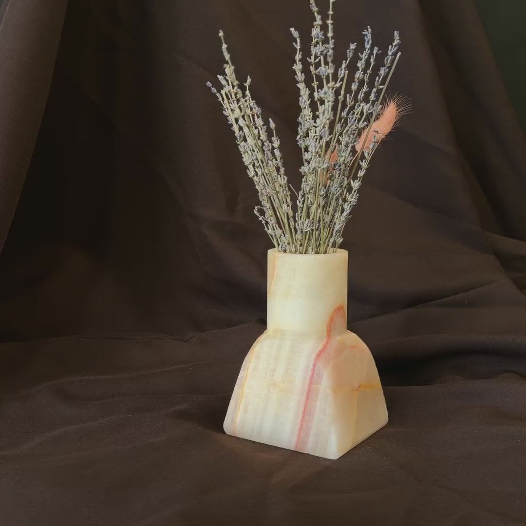 Onyx dune vase turning slowly to show its coral seam and milk-white stone, holding dried lavender and pink bunny tail grass against dark fabric