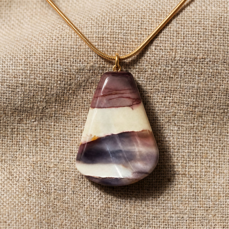 Teardrop-shaped multicolored stone pendant on a gold chain against a beige fabric background mookaite purple jasper