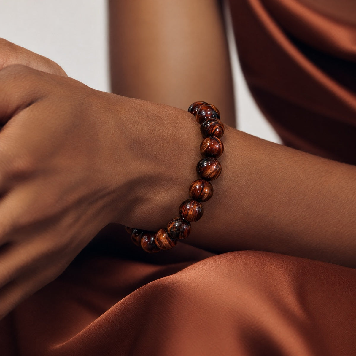 Red tiger's eye beaded stretch bracelet worn on a crossed wrist against rust-colored fabric.
