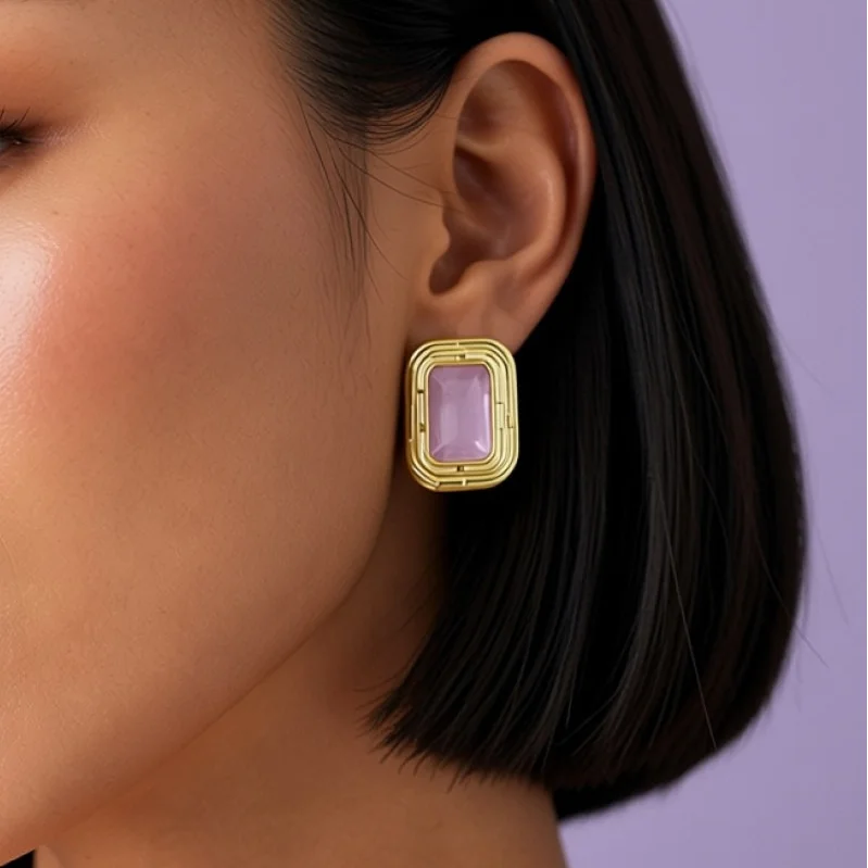Close detail of the rose cat's eye stud earring in its stepped gold frame, worn on the ear of a model with a dark bob, against a lilac background.