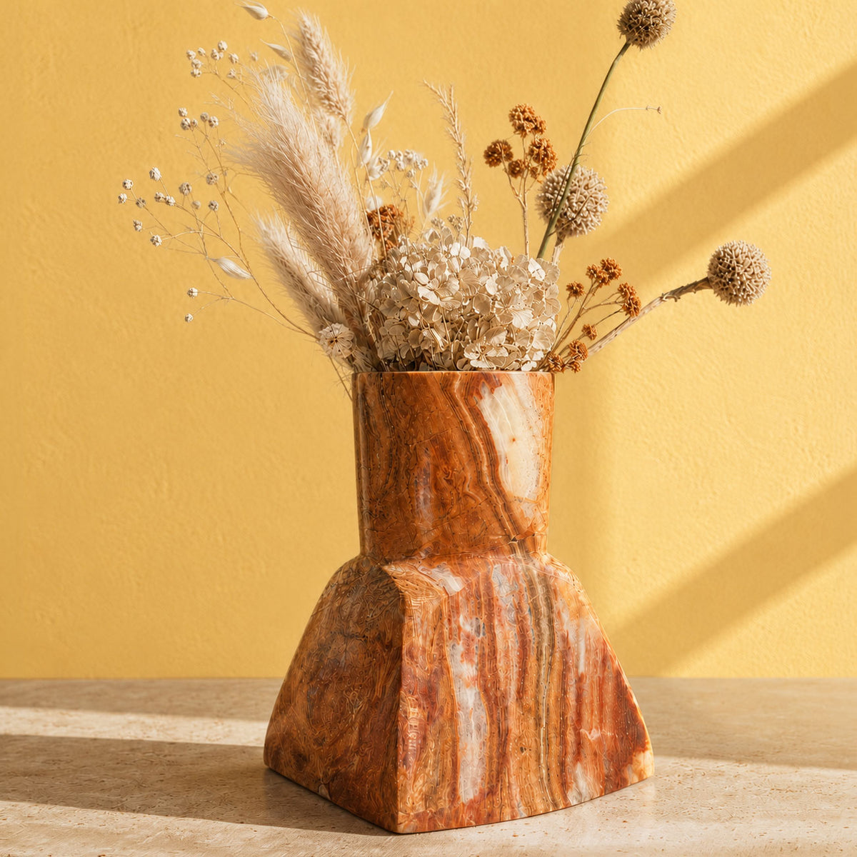 A sculptural terracotta-colored stone onyx vase with a flared base, filled with dried pampas grass and small white flowers, set against a warm yellow wall with soft diagonal shadows.