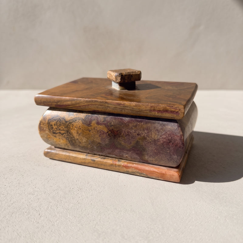 Rust and violet onyx keepsake box with lid in a three-quarter front view: a bulged body clouded in violet, honey gold and rust beneath a brown lid, a stepped base and a small square stone knob, in soft daylight