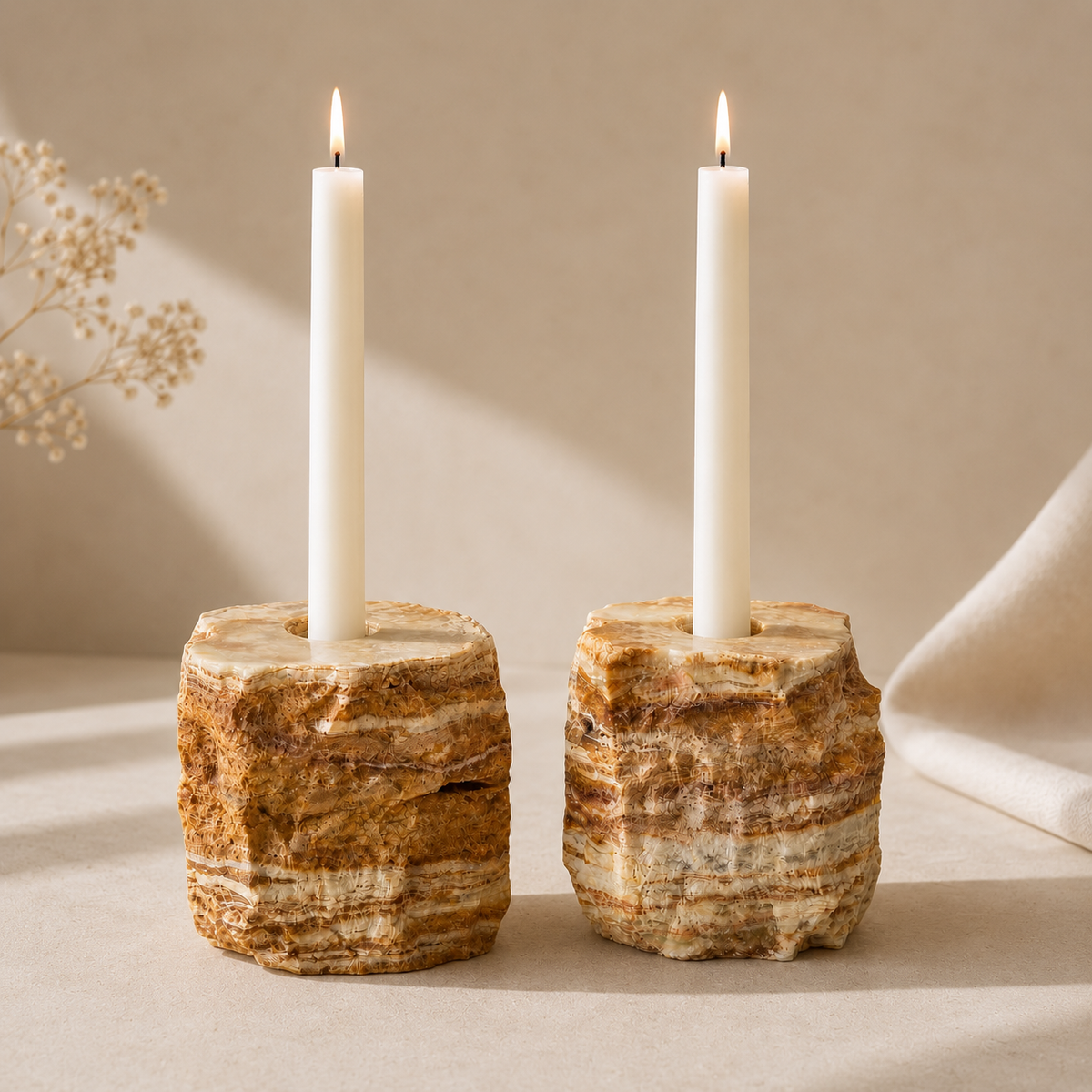 Two rustic onyx stone candle holders with white taper candles, styled on a neutral beige surface with soft shadows and dried flowers in the background.