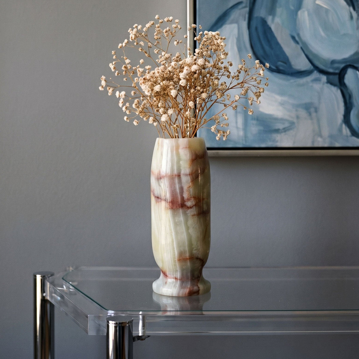 Decorative green light onyx vase with dried flowers on a glass table against a gray wall with abstract art.