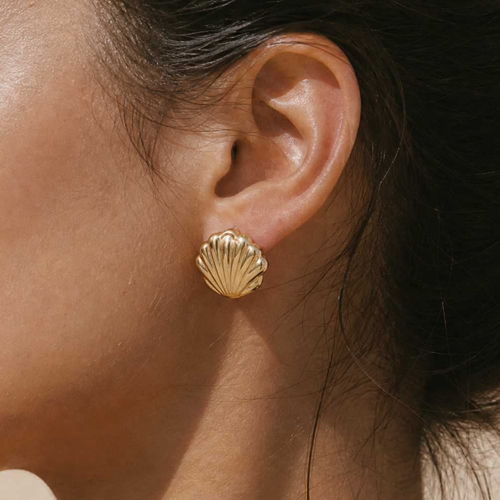 A close-up side profile of a woman wearing a small gold shell-shaped stud earring. The earring has a textured, ribbed surface and is shown against her skin and dark hair.