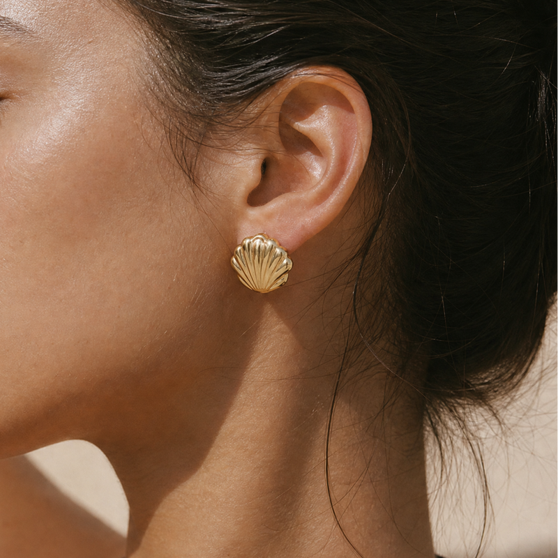 A close-up side profile of a woman wearing a small gold shell-shaped stud earring. The earring has a textured, ribbed surface and is shown against her skin and dark hair.