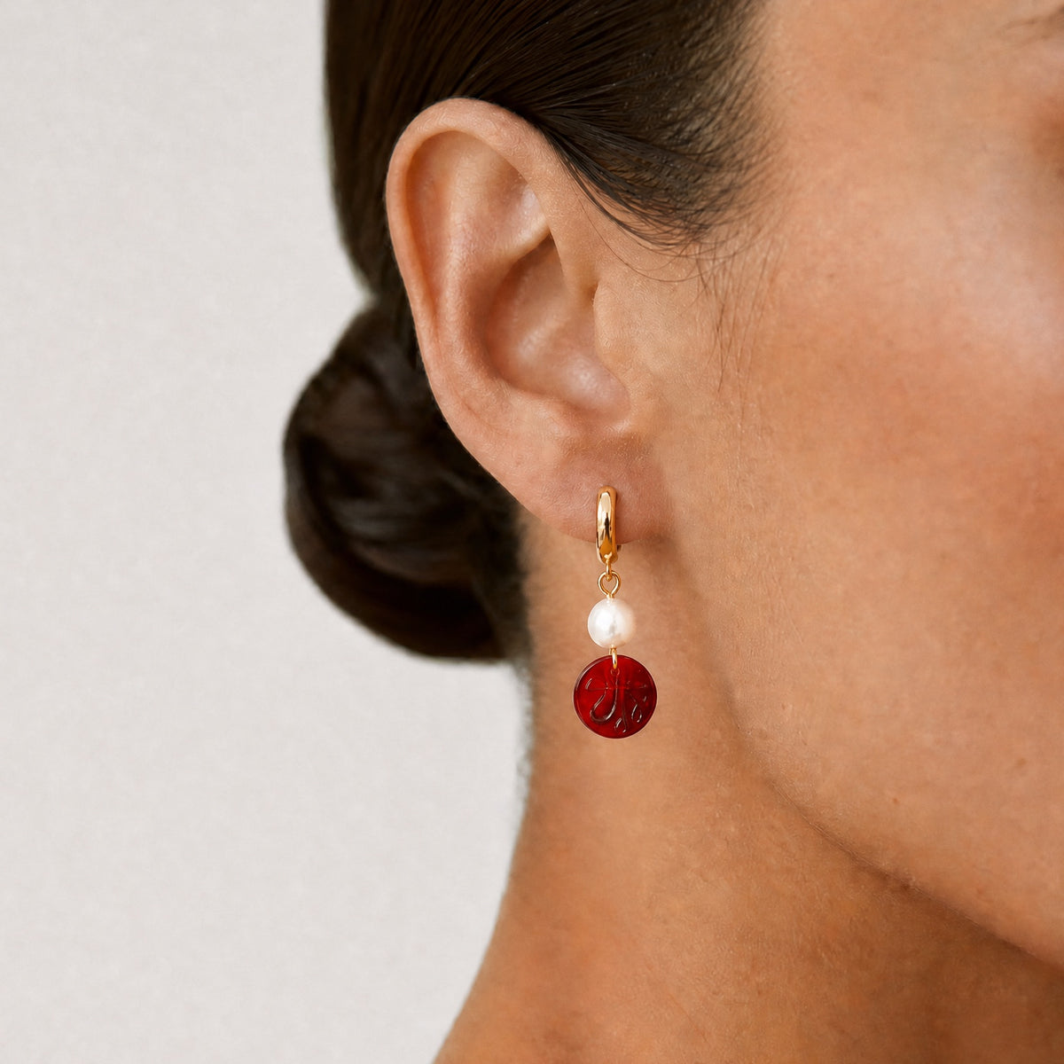 Close-up of a person wearing a red and gold hoop earring with an Allah sign on it on a plain background - Allah jewelry - Mecca beads - red garnet beads
