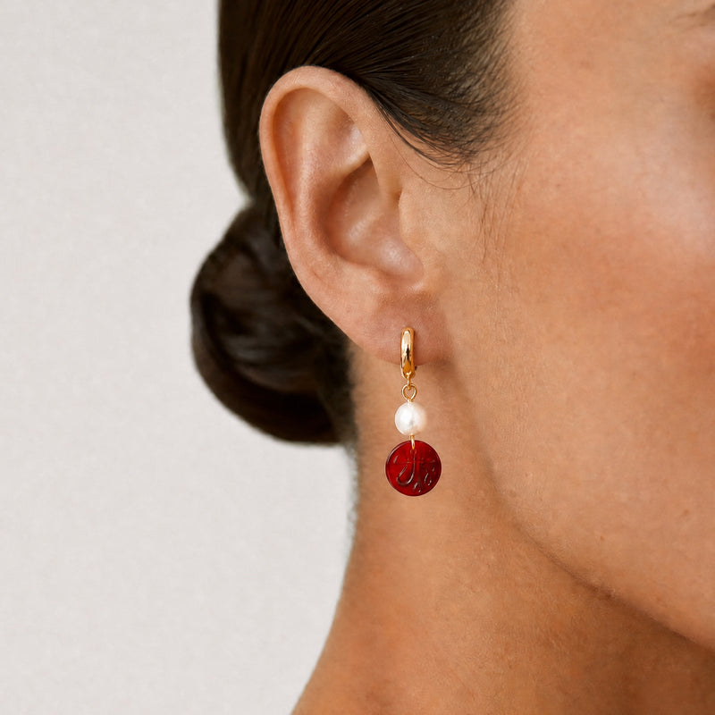 Close-up of a person wearing a red and gold hoop earring with an Allah sign on it on a plain background - Allah jewelry - Mecca beads - red garnet beads