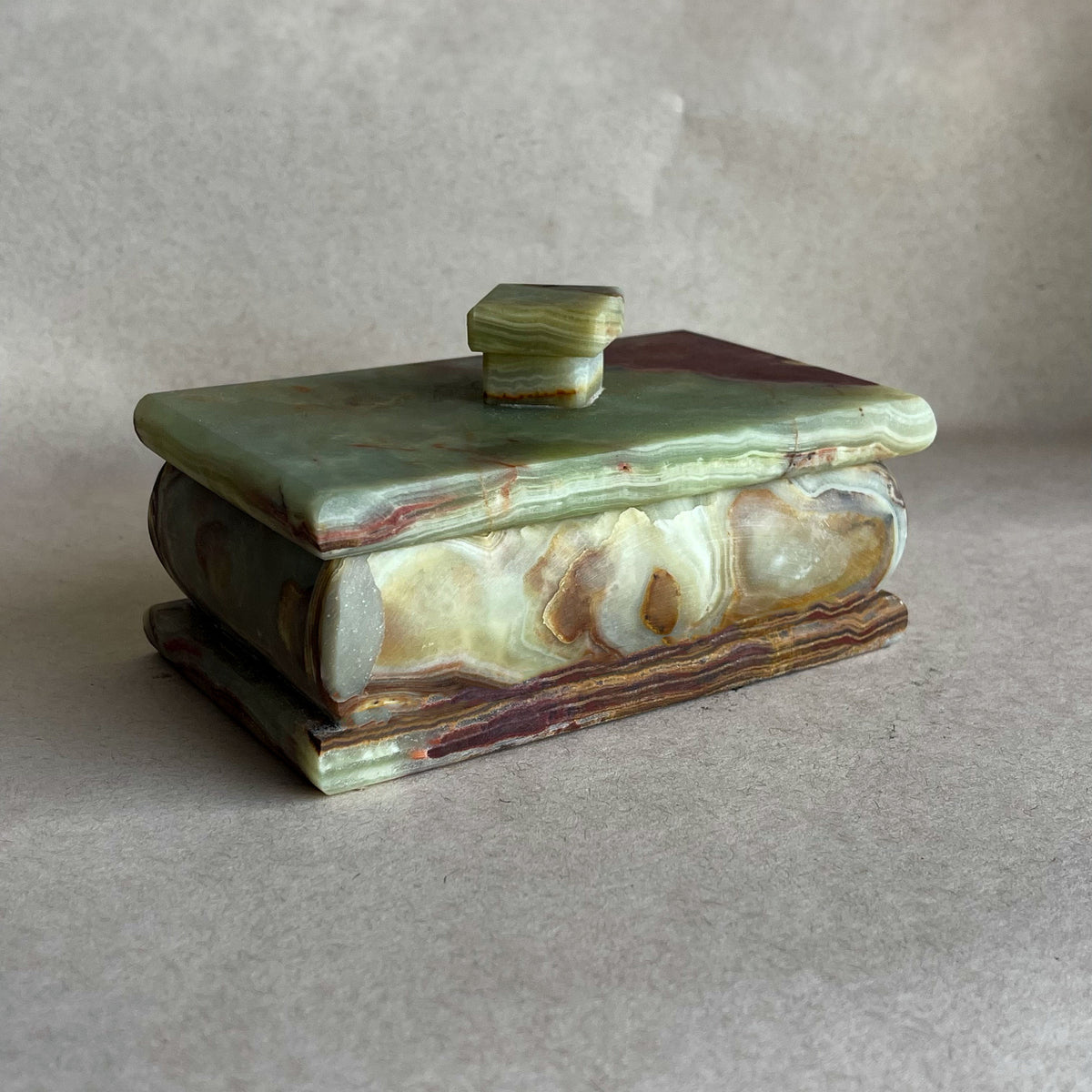 Hand-carved sage green and rust onyx lidded keepsake box with sculptural knob, showing natural jade, cream, and amber striations on neutral backdrop
