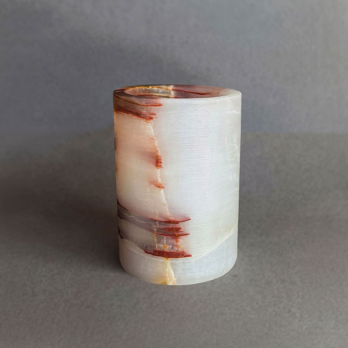 hand-carved natural onyx vessel in cream and rust tones with bold amber veining, cylindrical stone holder on gray background.