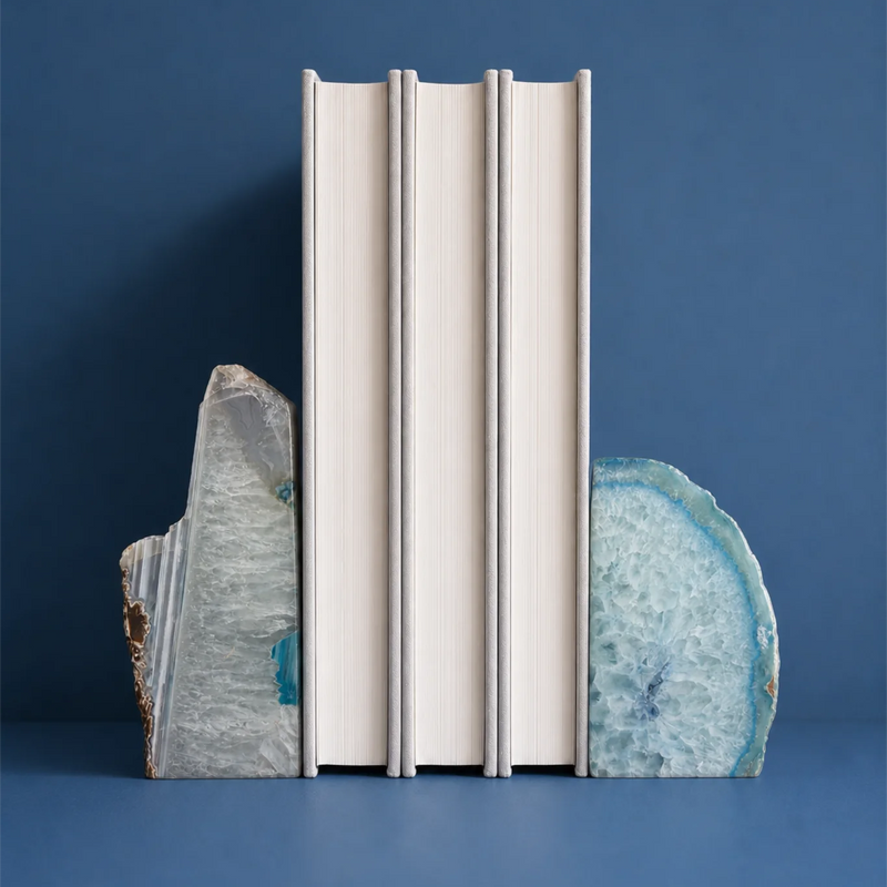 Three slim off-white books standing upright between two polished agate slice bookends on a blue surface and background.